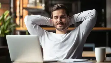 Man with hands behind head looking at a laptop screen.